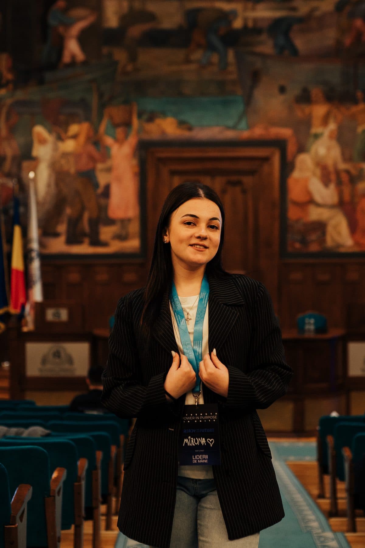 Voluntară zâmbitoare în sacou negru, cu ecuson, în aula ASE cu pictura murală în spate