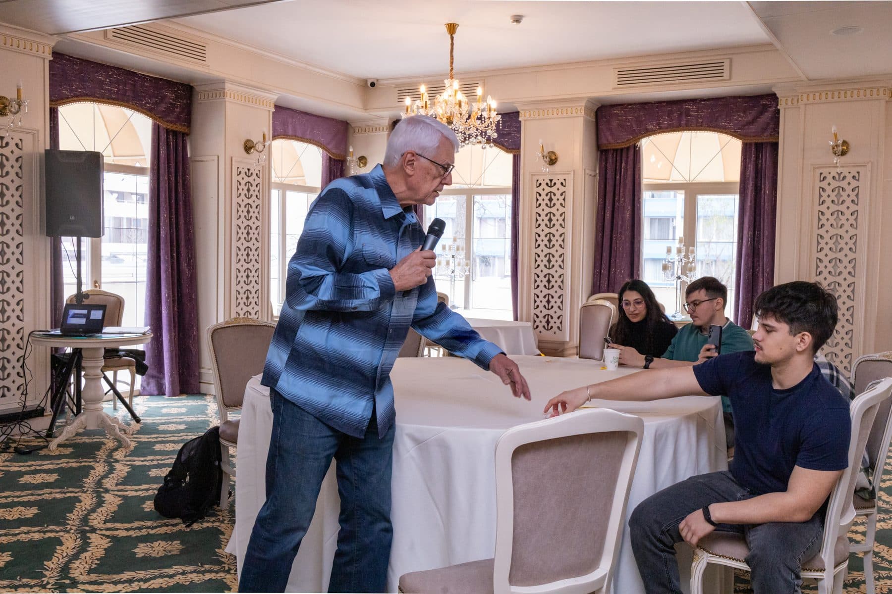 Mentor vârstnic cu microfon se sprijină de o masă și vorbește cu studenții așezați la ea, la teambuilding