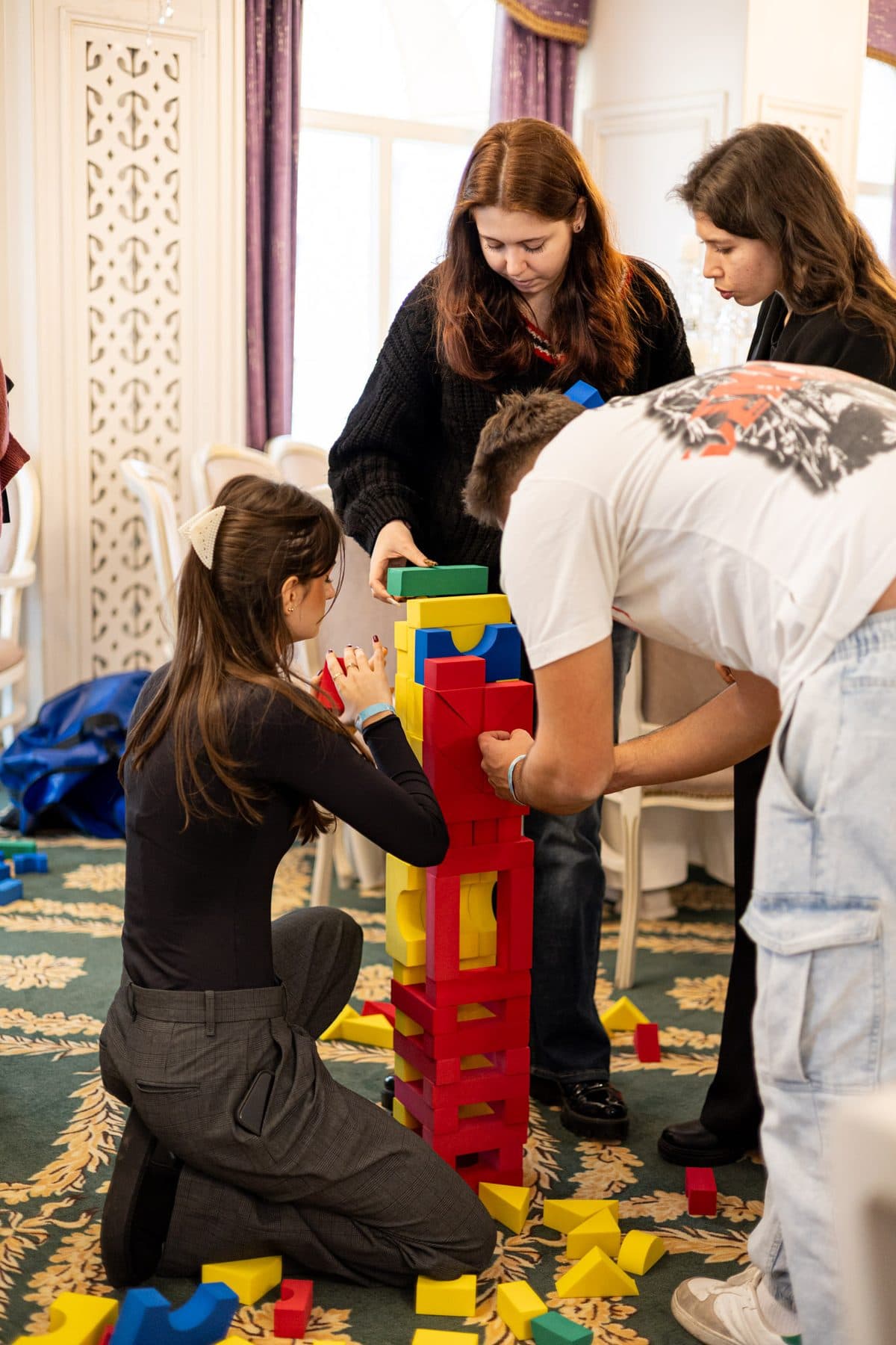 Patru studenți construiesc împreună un turn înalt din cuburi roșii și galbene, pe covorul sălii de teambuilding