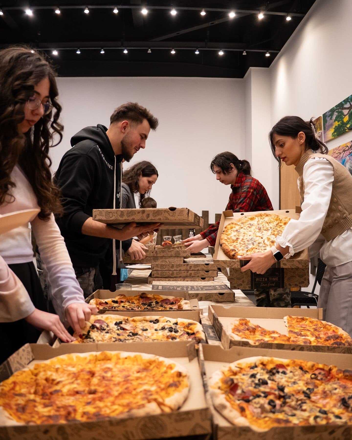 Masă lungă cu cutii de pizza deschise, în jurul căreia se strâng studenții după întâlnire