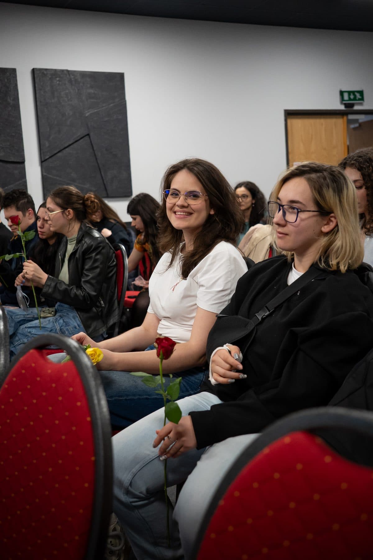 Două studente zâmbitoare țin în mână trandafiri primiți la întâlnirea din martie, așezate pe scaune roșii