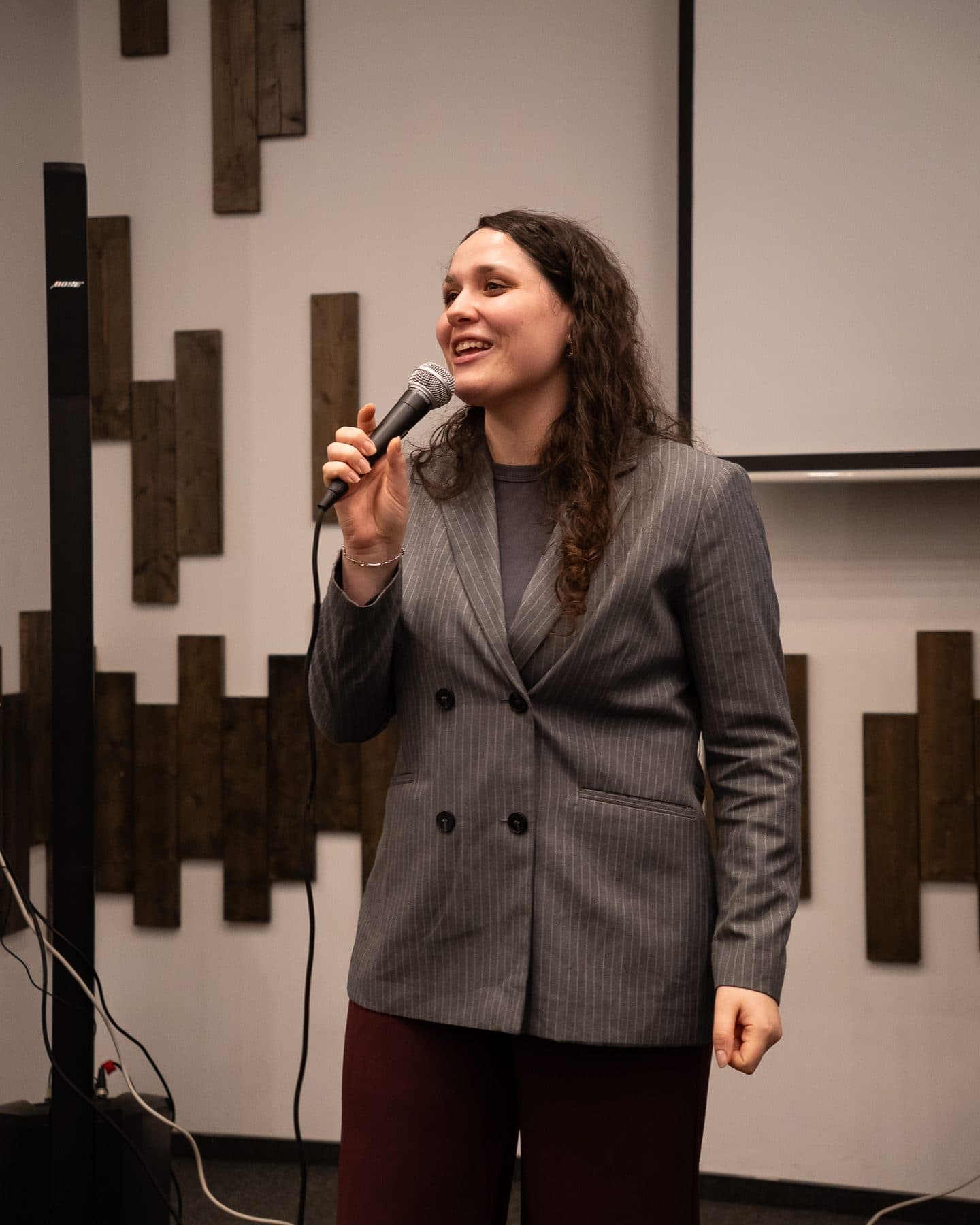 Tânără în sacou gri vorbește zâmbind la microfon, în fața unui perete cu lambriuri de lemn