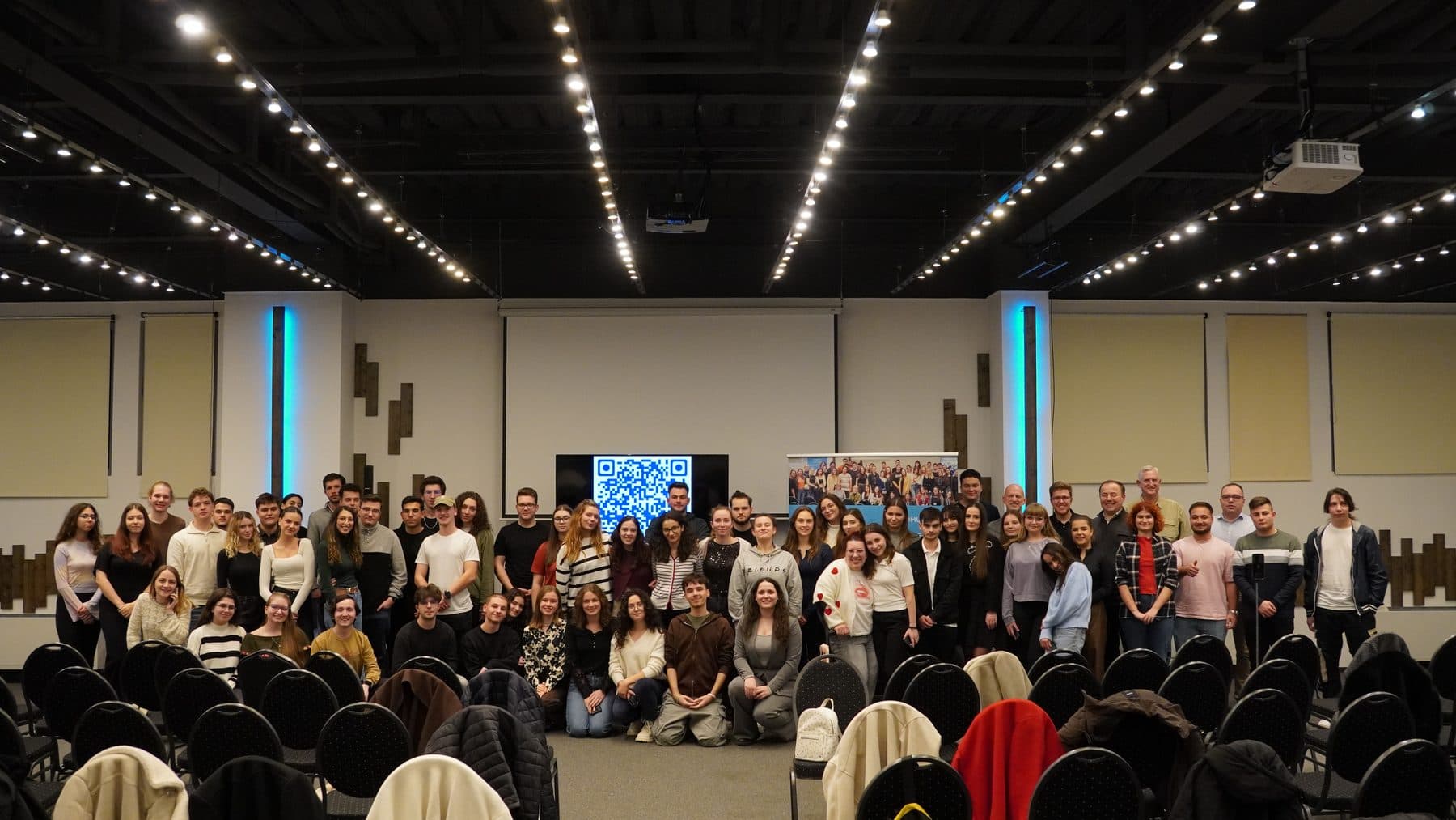 Zeci de studenți Liderii de Mâine pozează împreună în fața unei săli mari de conferințe, sub lumini în formă de stele