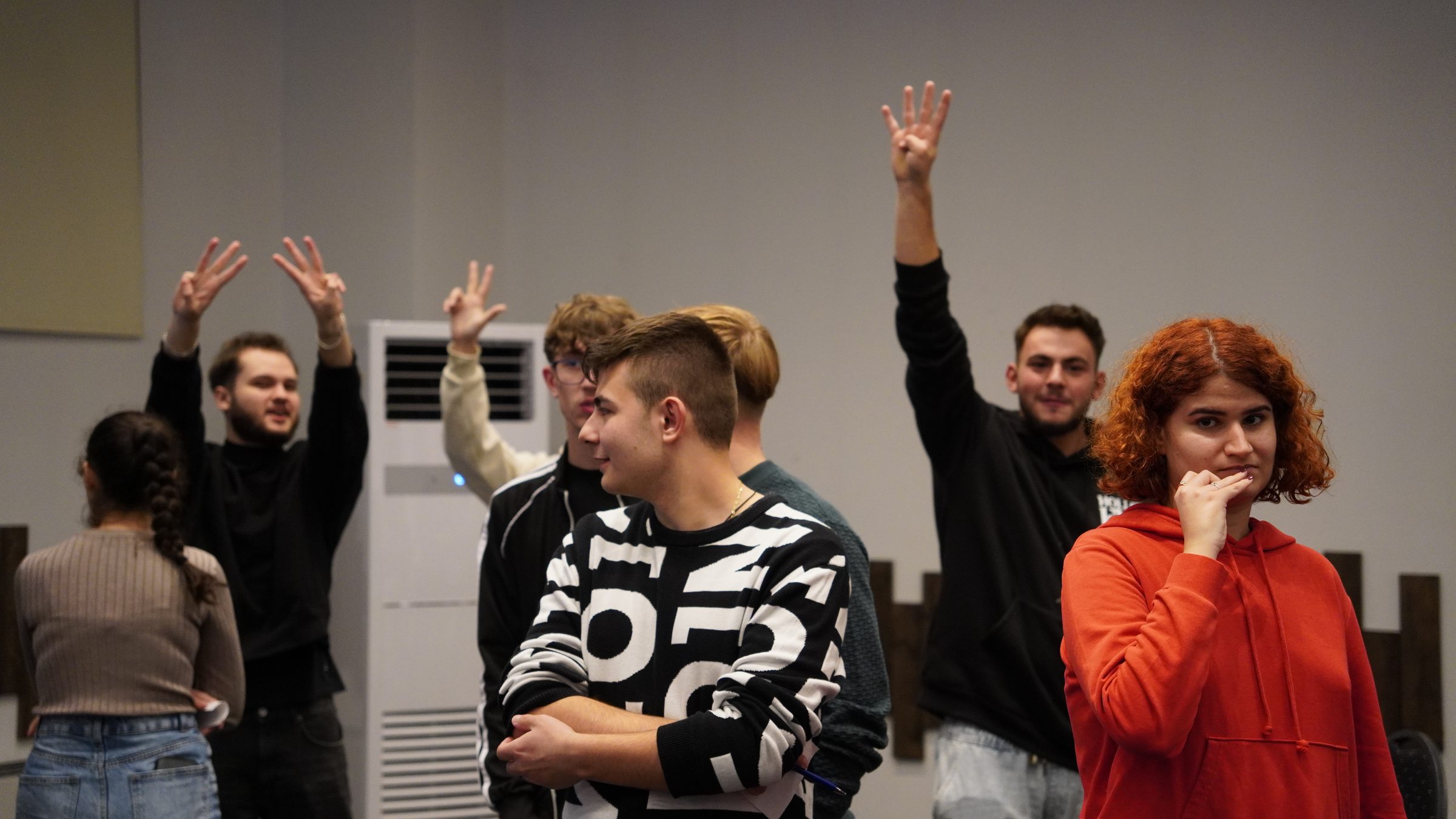 Studenți care ridică mâinile cu entuziasm în timpul unui joc la întâlnirea de marți, o tânără roșcată zâmbind în prim-plan