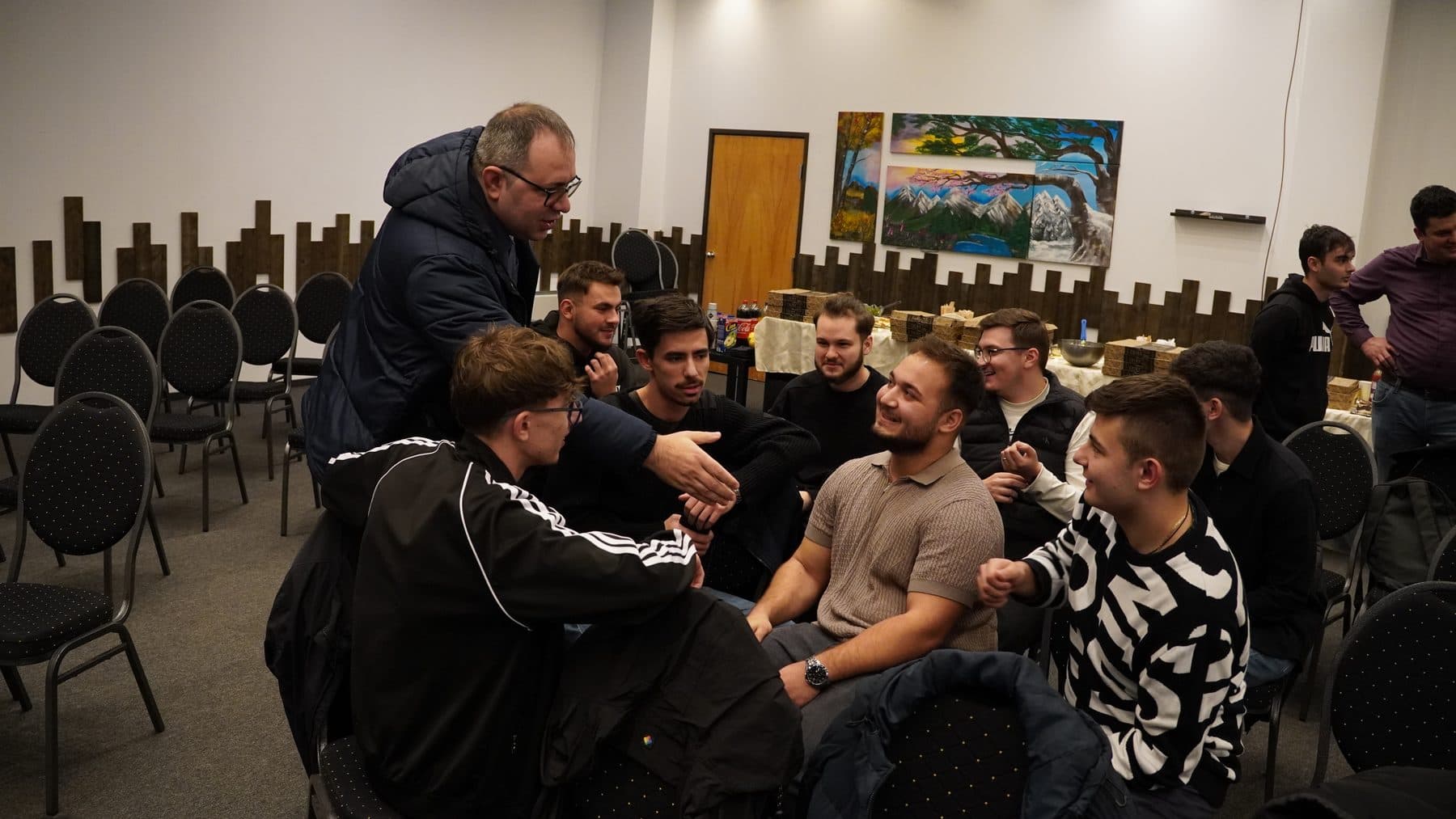 Mentor aplecat spre un cerc de tineri așezați, care zâmbesc și discută cu el la întâlnirea de marți