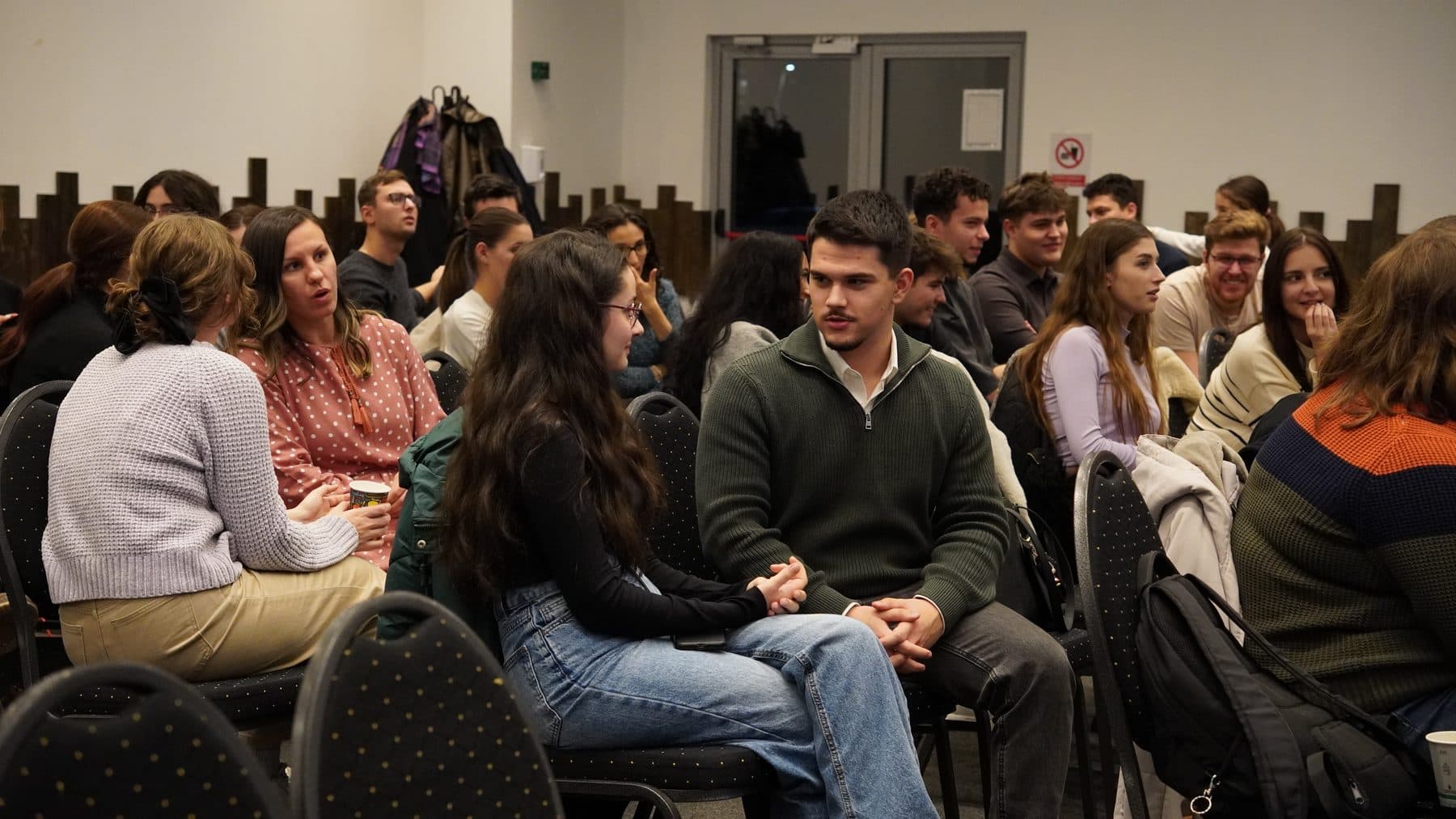 Sală plină de studenți așezați față în față, discutând în perechi și grupuri mici la întâlnirea de marți