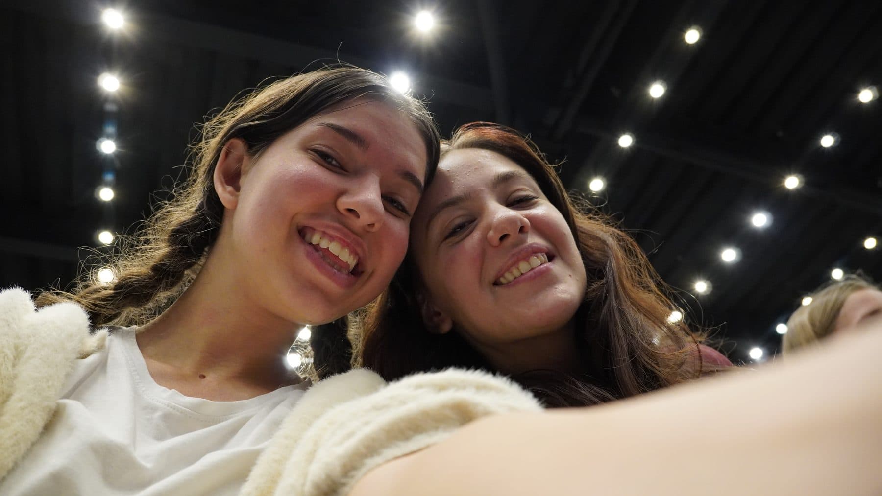 Două prietene își fac un selfie zâmbind sub luminile sălii de întâlnire
